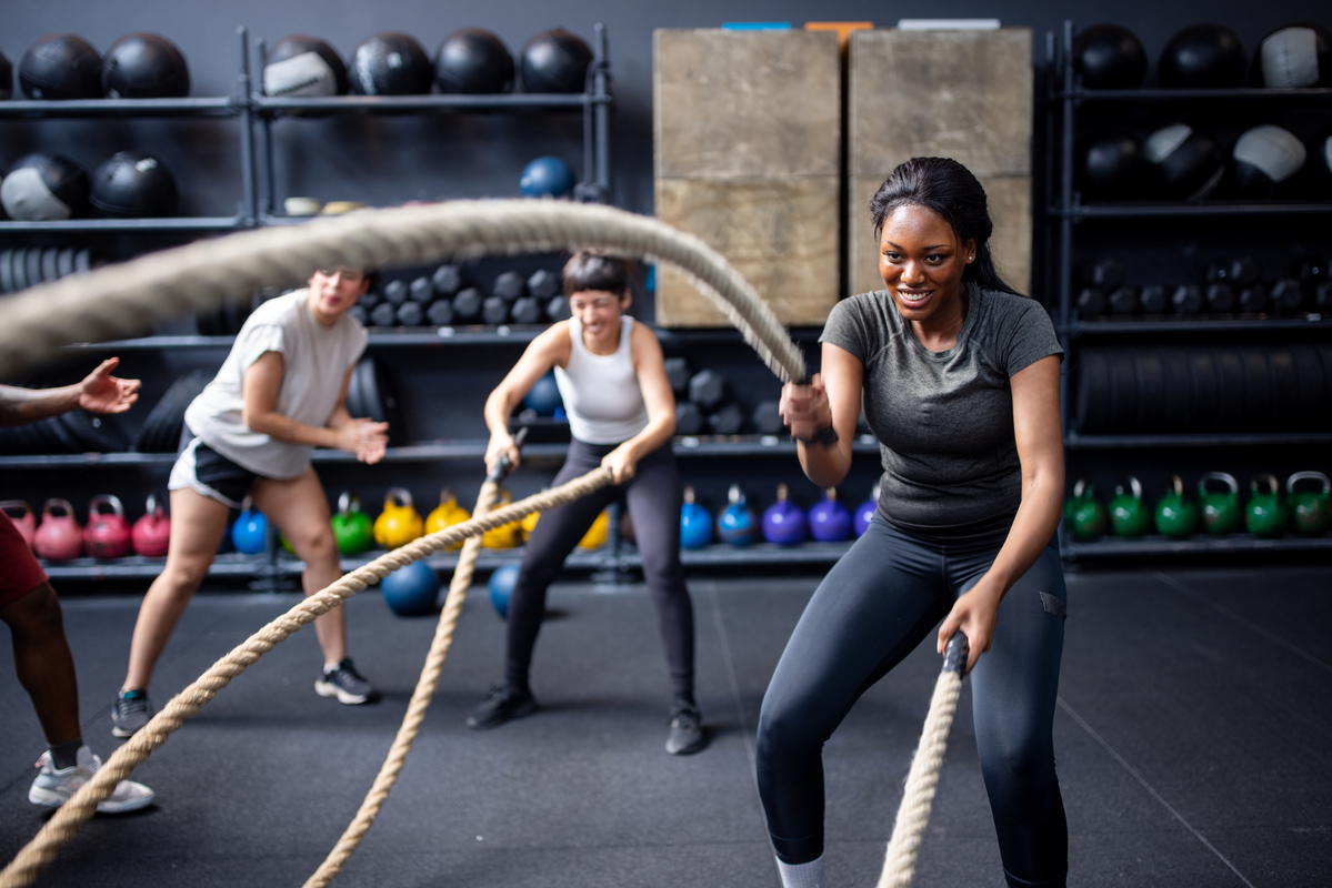 Two people in a gym, working out with battle ropes while a friend cheers them on.