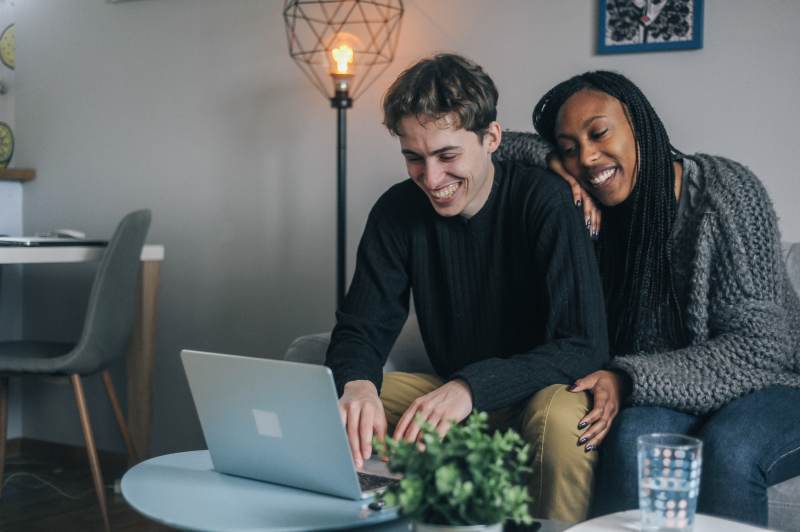Man using laptop to apply for a job, partner leaning on his shoulder smiling.