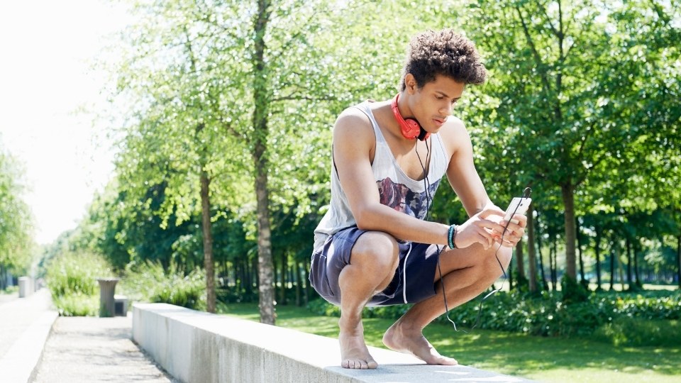 A young man wearing headphones around his neck crouches on a cement ledge as he checks his phone.