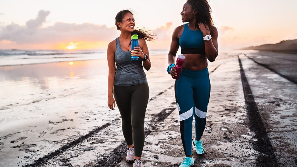 Two female friends walk along the beach at sunset while holding drink bottles 