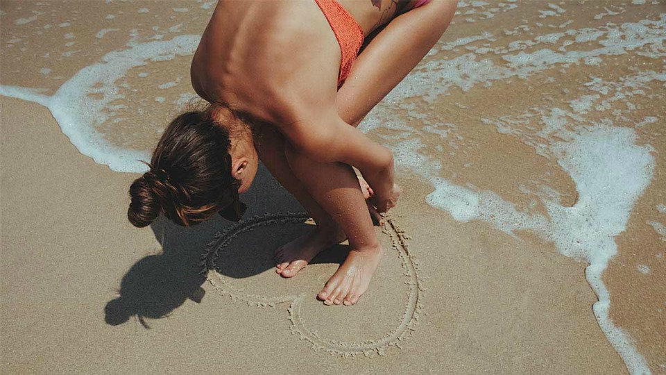 Person standing in the middle of a love-heart shape drawn in the sand at the beach.