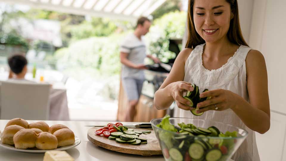 A woman prepares a salad as her husband cooks a BBQ and their son sits at the table.