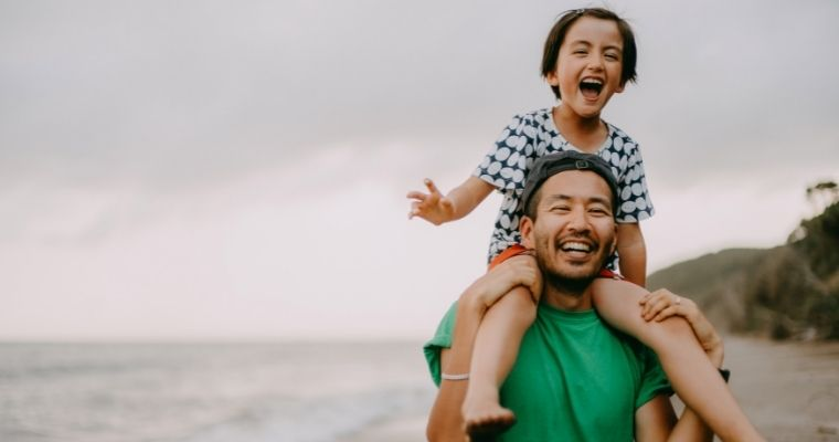 A child sitting on an adults shoulders, they are walking along the beach and both are laughing