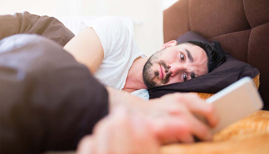 An unwell young man laying in bed while looking at his phone.