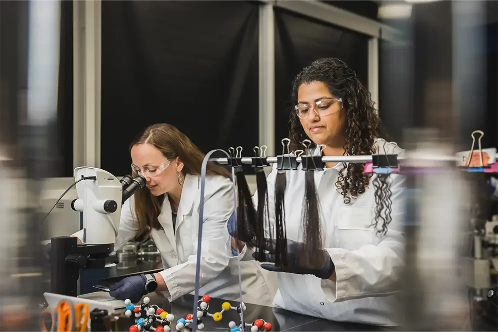 Két laboratóriumi dolgozó hajmintákat elemez és anyagokat vizsgál mikroszkóppal.