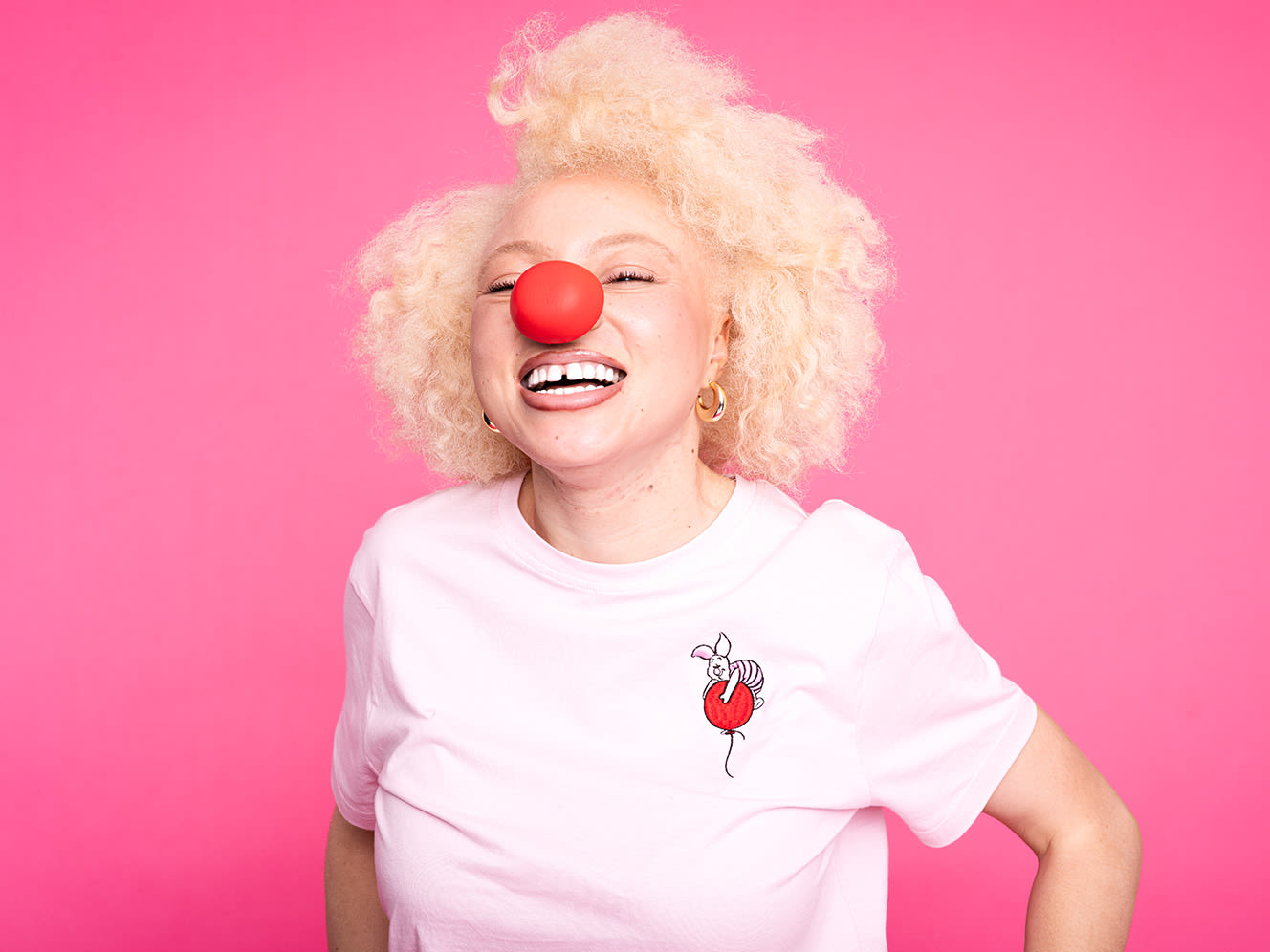 Smiling person with blonde hair wears a red nose and pink shirt, against a bright pink background