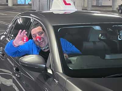 A driving instructor sits in their car and wears a red nose for Comic Relief
