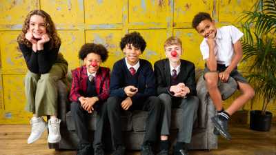 Five young people sitting on a sofa smiling, two individuals wear Comic Relief red noses.