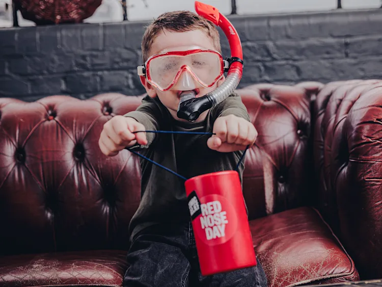 A boy smiling and wearing a red scuba mask holds a Red Nose Day coin collector.