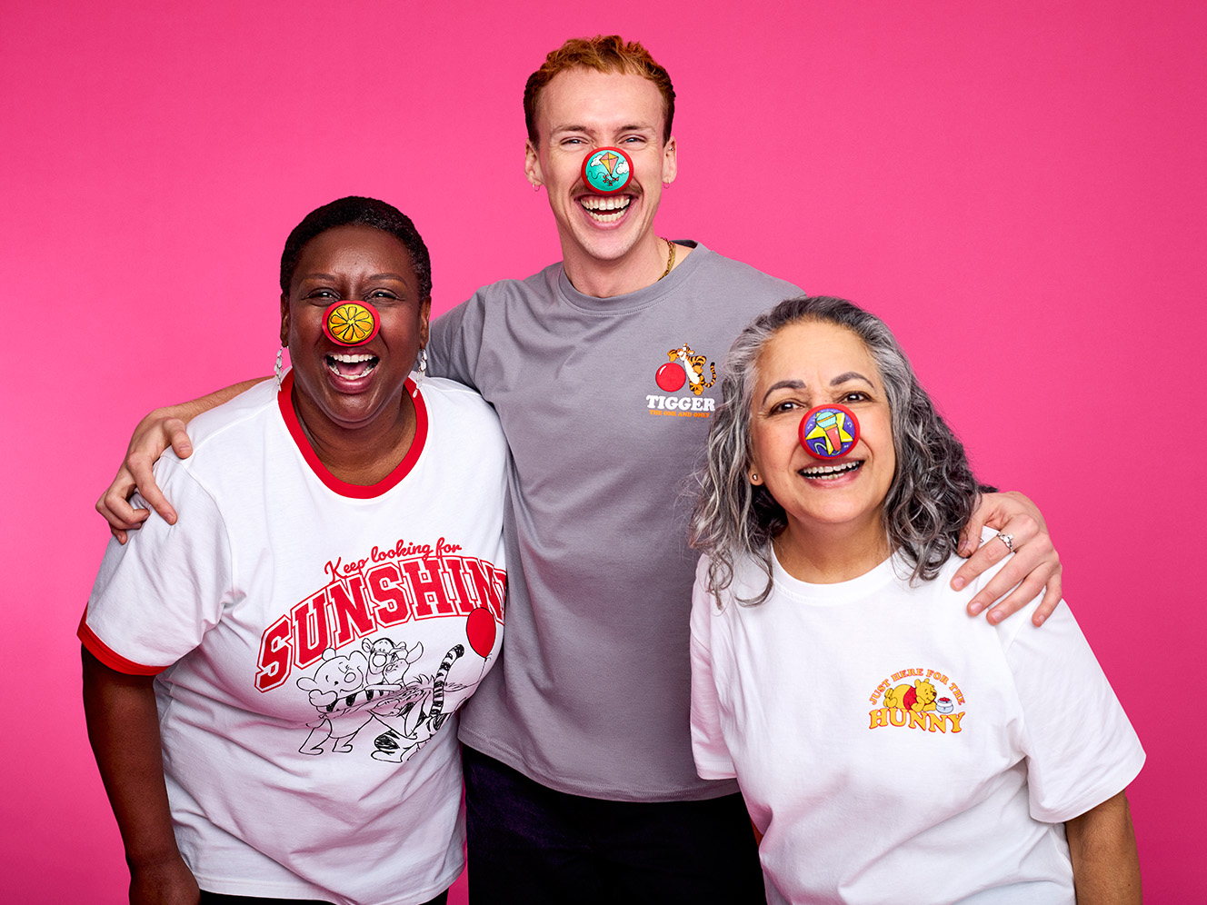 Three individuals pose together, wearing fun, themed shirts against a bright pink background, showcasing a cheerful and playful atmosphere