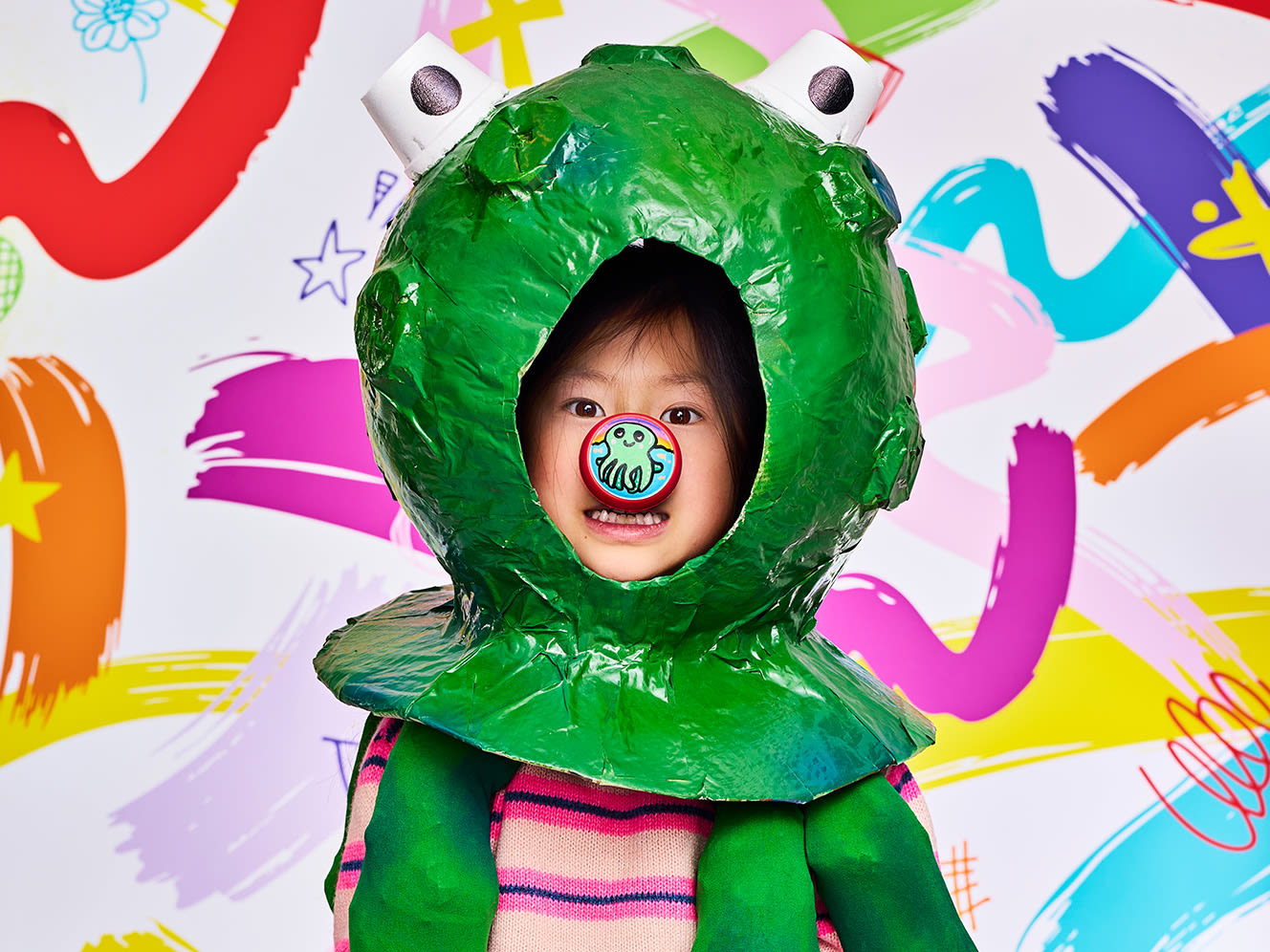 A child wearing a green monster costume with eye stalks standing against a colourful, abstract background, sports a cartoon hand-painted red nose