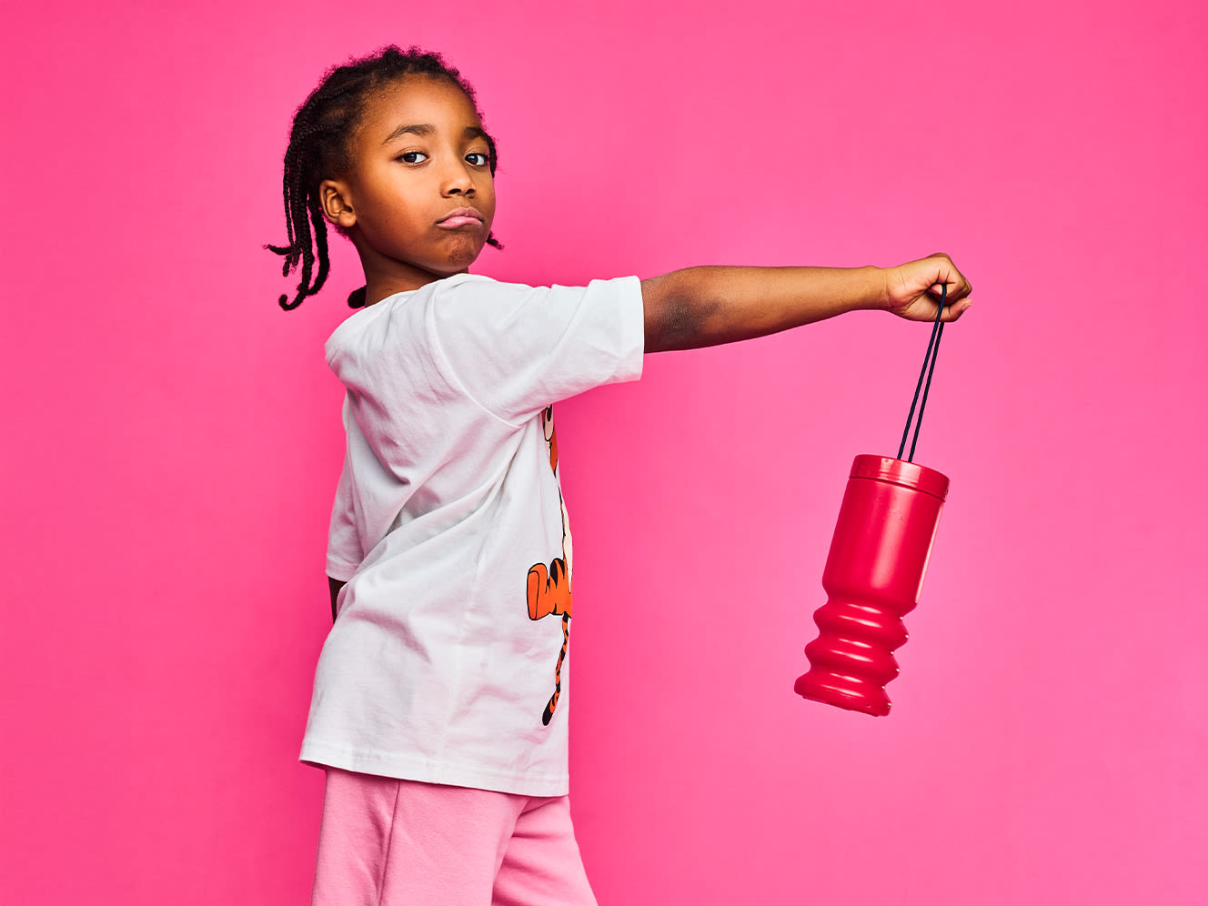 A young child confidently holds a red money box against a vibrant pink background