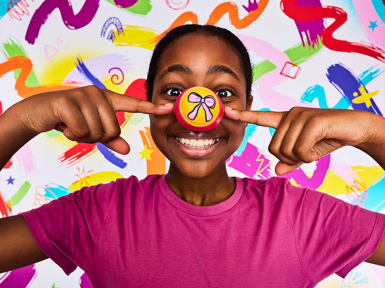 A child smiles, pointing to a hand-painted red nose with a pink bow design. Set against a vibrant abstract background.