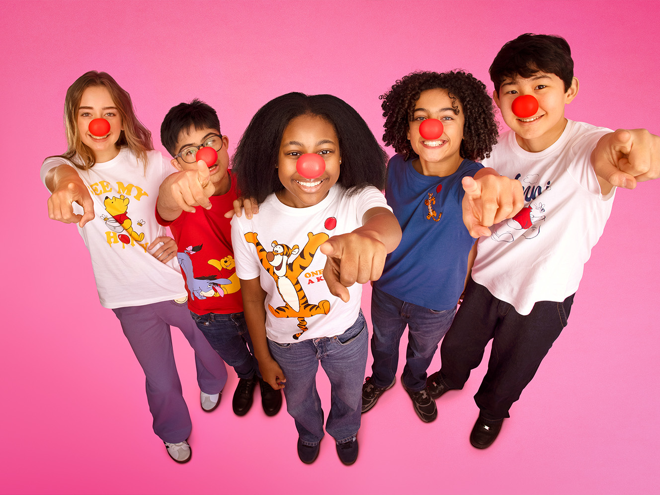 Five children with red noses happily point at the camera against a bright pink background