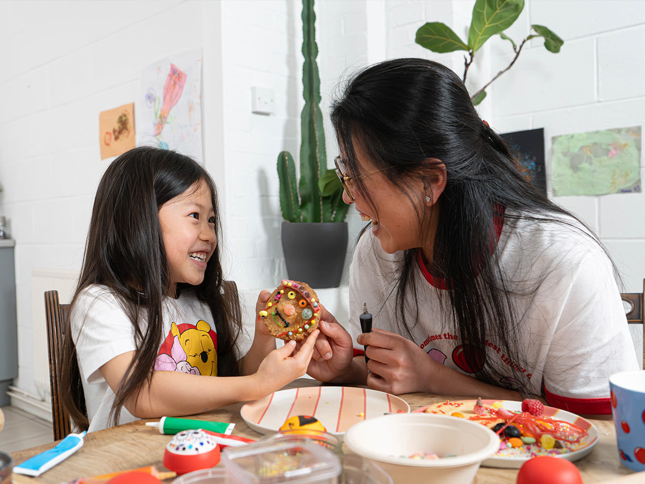 Mother and daughter smiling a each other while decorating a cookie at a table with colourful treats
