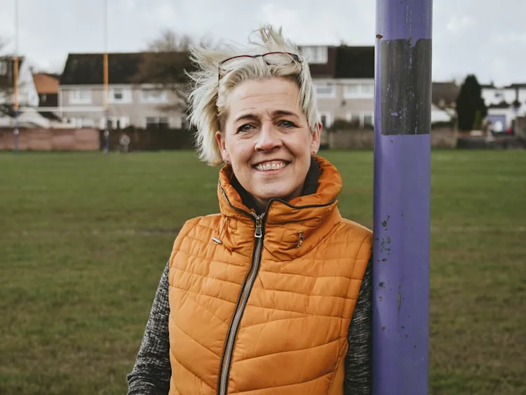 Kate leans against a rugby goal post and smiles at the camera.
