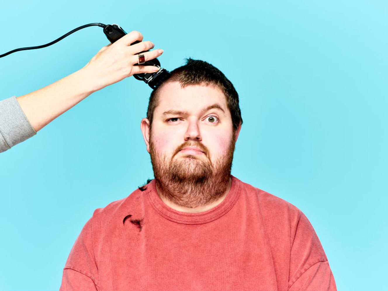 A man having his hair clipped, wearing a red shirt and has a humorous, uncertain expression against blue background