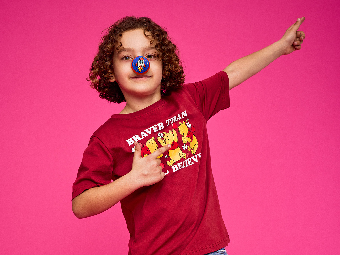 Child with curly hair poses confidently against a bright pink background, wearing a red nose and a red shirt, pointing playfully