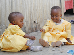 Two infants sitting on the floor, wearing yellow dresses