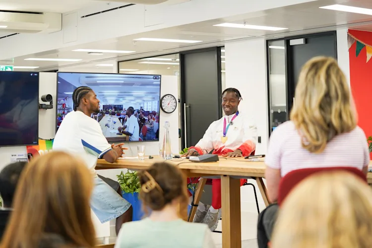 Two speakers in conversation at a Comic Relief staff event, with colleagues watching in the office and joining on screen