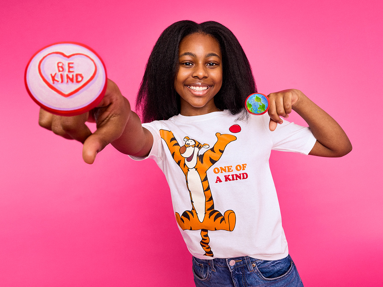 A smiling young girl in a Tigger shirt, pointing at the camera, holds decorative hand-painted red noses, one with "BE KIND" in a heart, and another of the Earth. Standing against a bright pink background