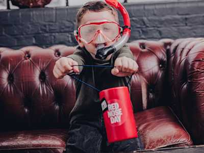 A boy smiling and wearing a red scuba mask holds a Red Nose Day coin collector.