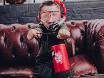 A boy smiling and wearing a red scuba mask holds a Red Nose Day coin collector.