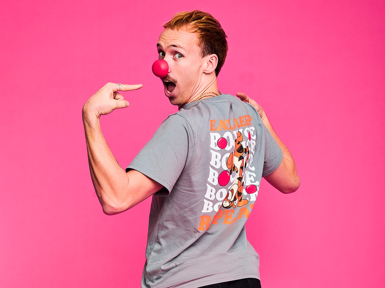 Young person in a grey printed shirt, wearing a red nose, playfully poses against a bright pink background