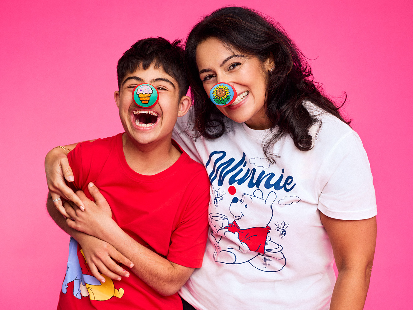 A joyful woman and child, wearing decorative red noses, hug and laugh against a vibrant pink background