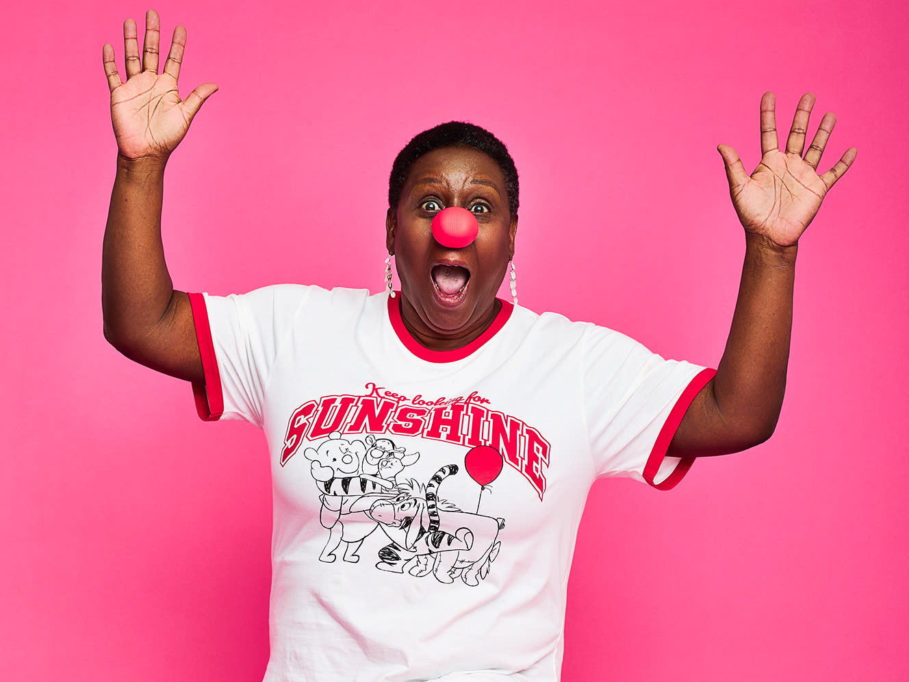 A person with a red nose and a surprised expression raises their hands against a bright pink background, exuding joy and playfulness