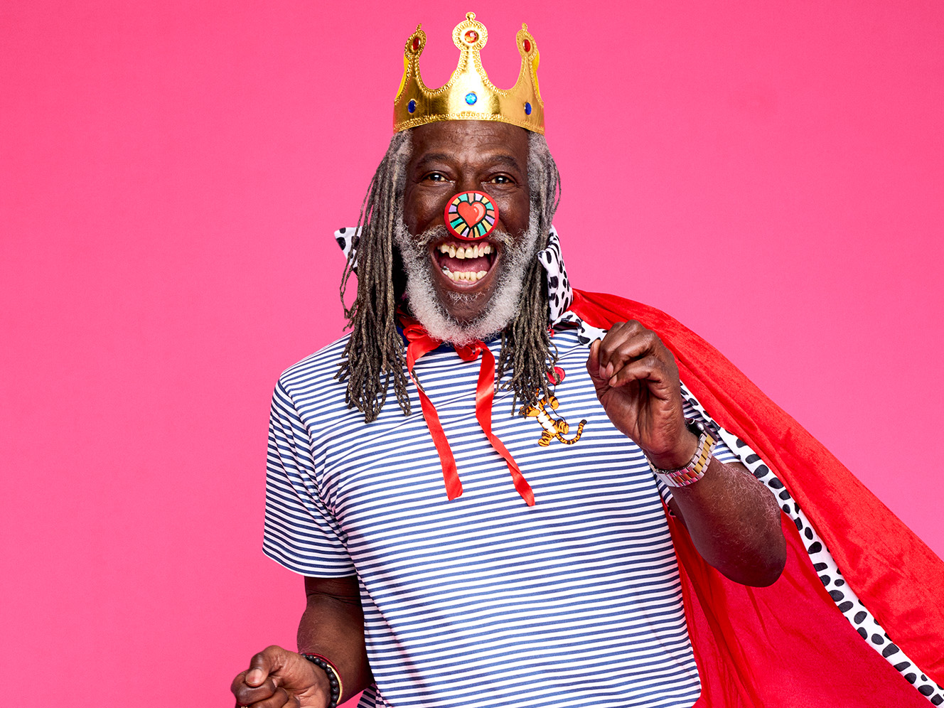 A man with a golden crown, red nose and cape, smiling joyfully against a bright pink background