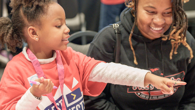 Child points at craft materials while smiling adult looks on at a table activity.