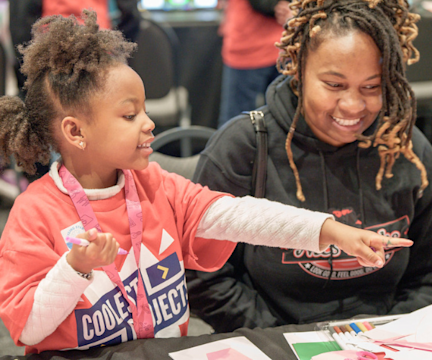 Child points at craft materials while smiling adult looks on at a table activity.