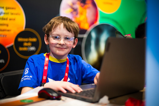 Child using a laptop at a coding event, wearing safety glasses and a Raspberry Pi Foundation T-shirt.