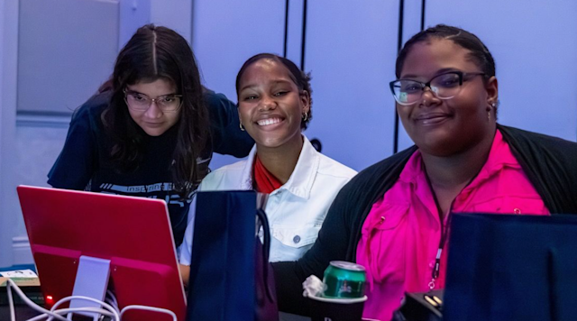 three people sit at a desk using laptops, one smiling at the camera while others focus on their screens.