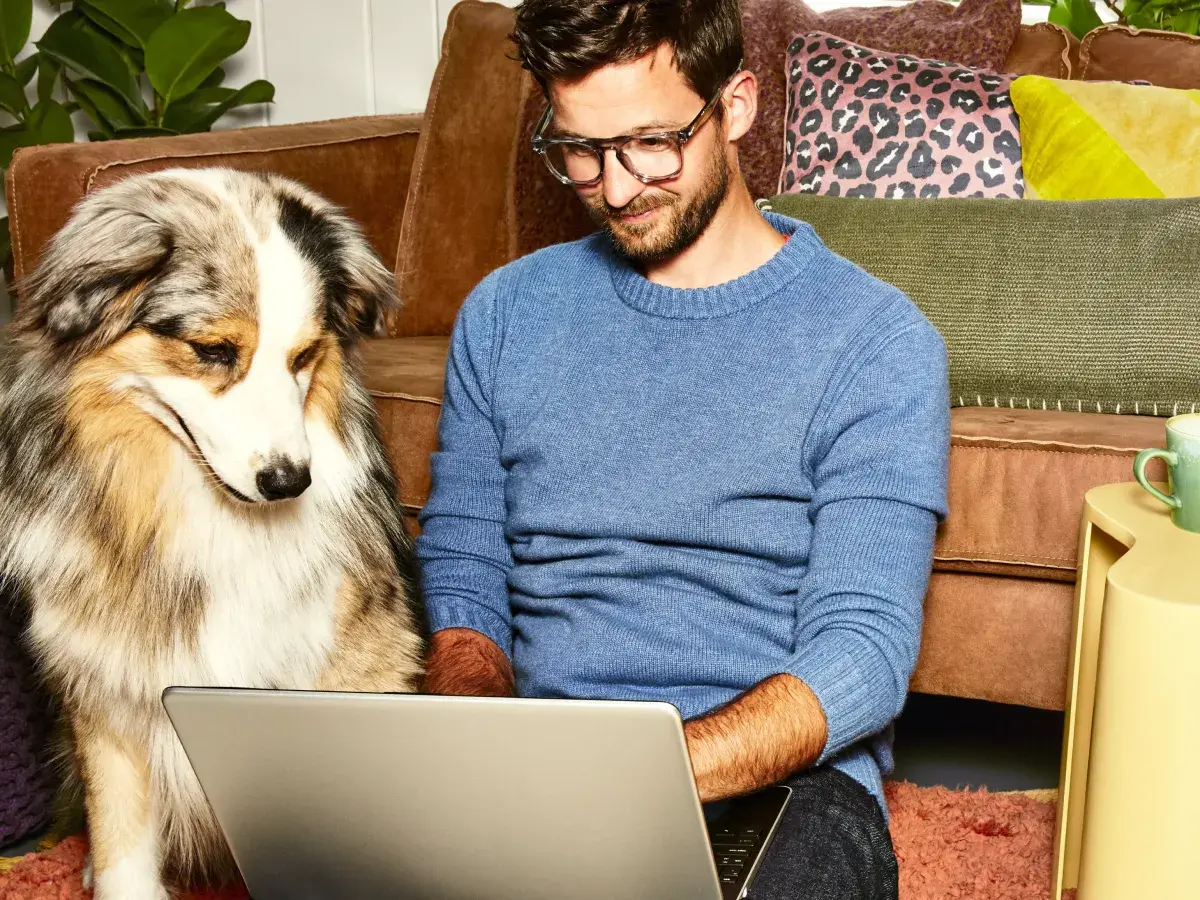 Man zit op de grond tegen bank met laptop op schoot en hond ernaast