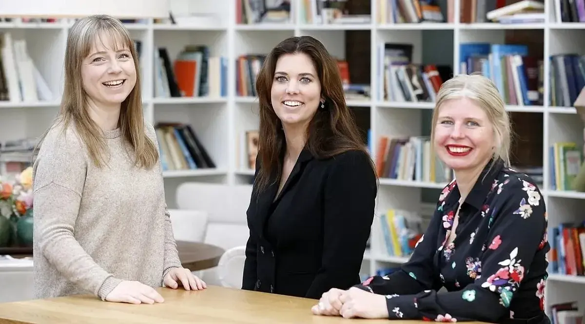 Drie dames staan in een bibliotheek  aan een tafel met elkaar te praten