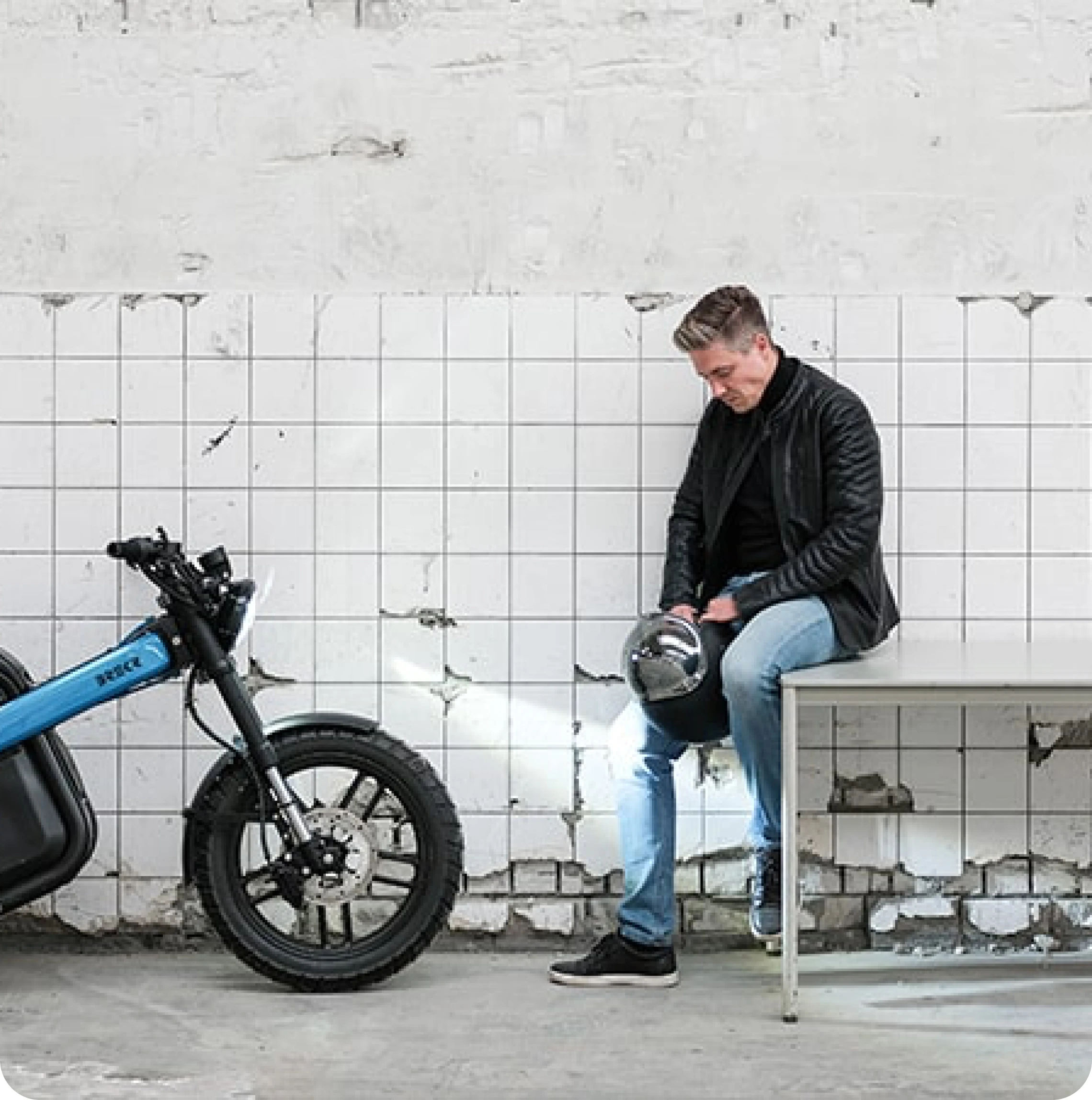 Meneer zit op een tafel. Naast hem staat een blauwe Brekr brommer 