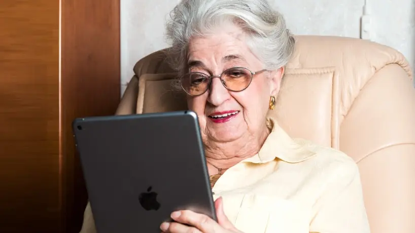 Een oudere dame zit op een stoel met een iPad in haar handen.