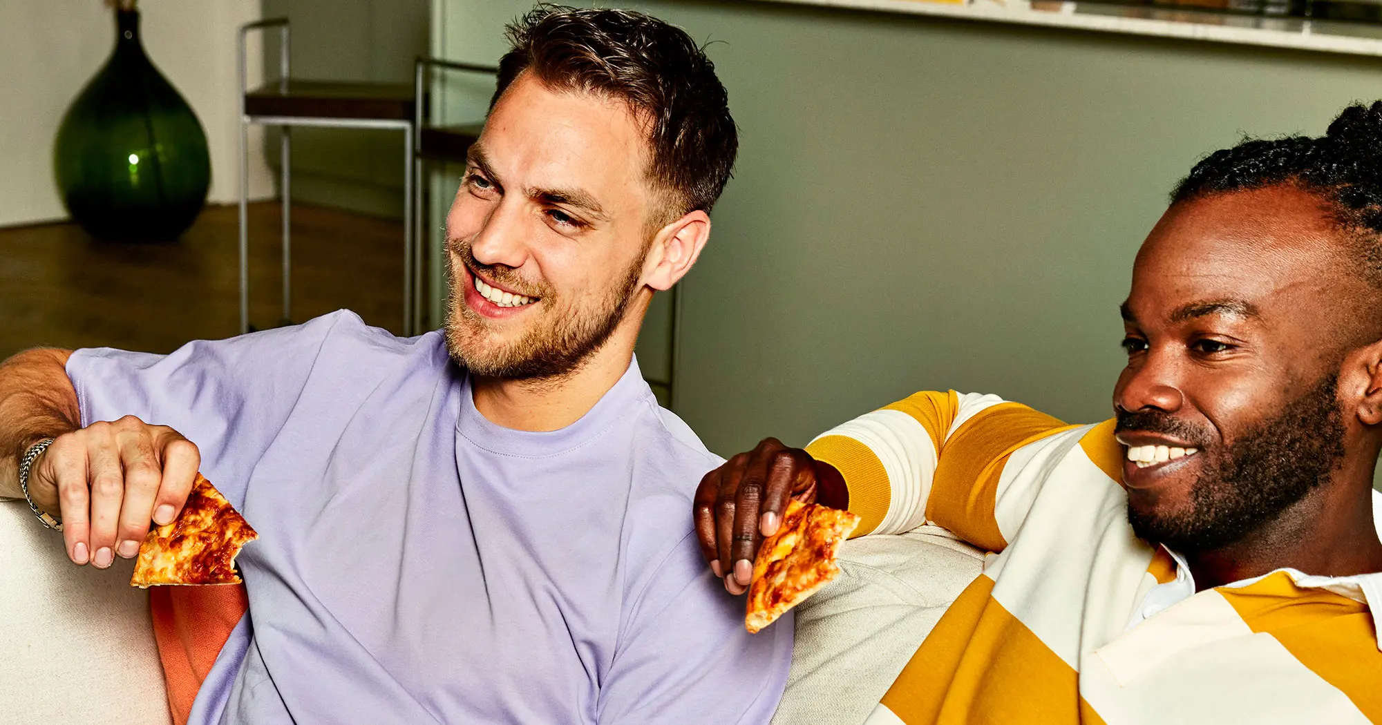 2 jongens kijken lachend tv met een stuk pizza in hun handen