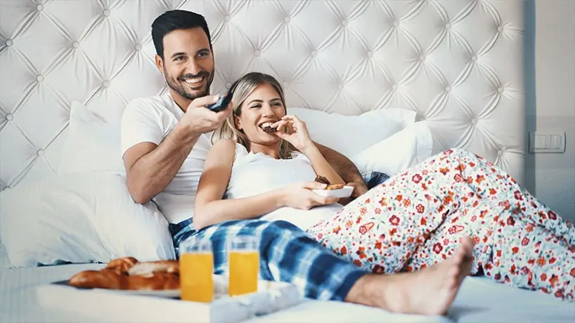 Een man en vrouw liggen op bed. Op het bed staat een dienblad met eten. De man en vrouw kijken tv.