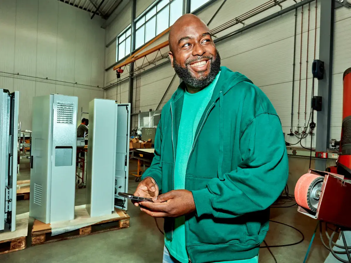 Een lachende man staat in een werkplaats. Hij heeft een groene jas aan en houdt zijn telefoon vast.