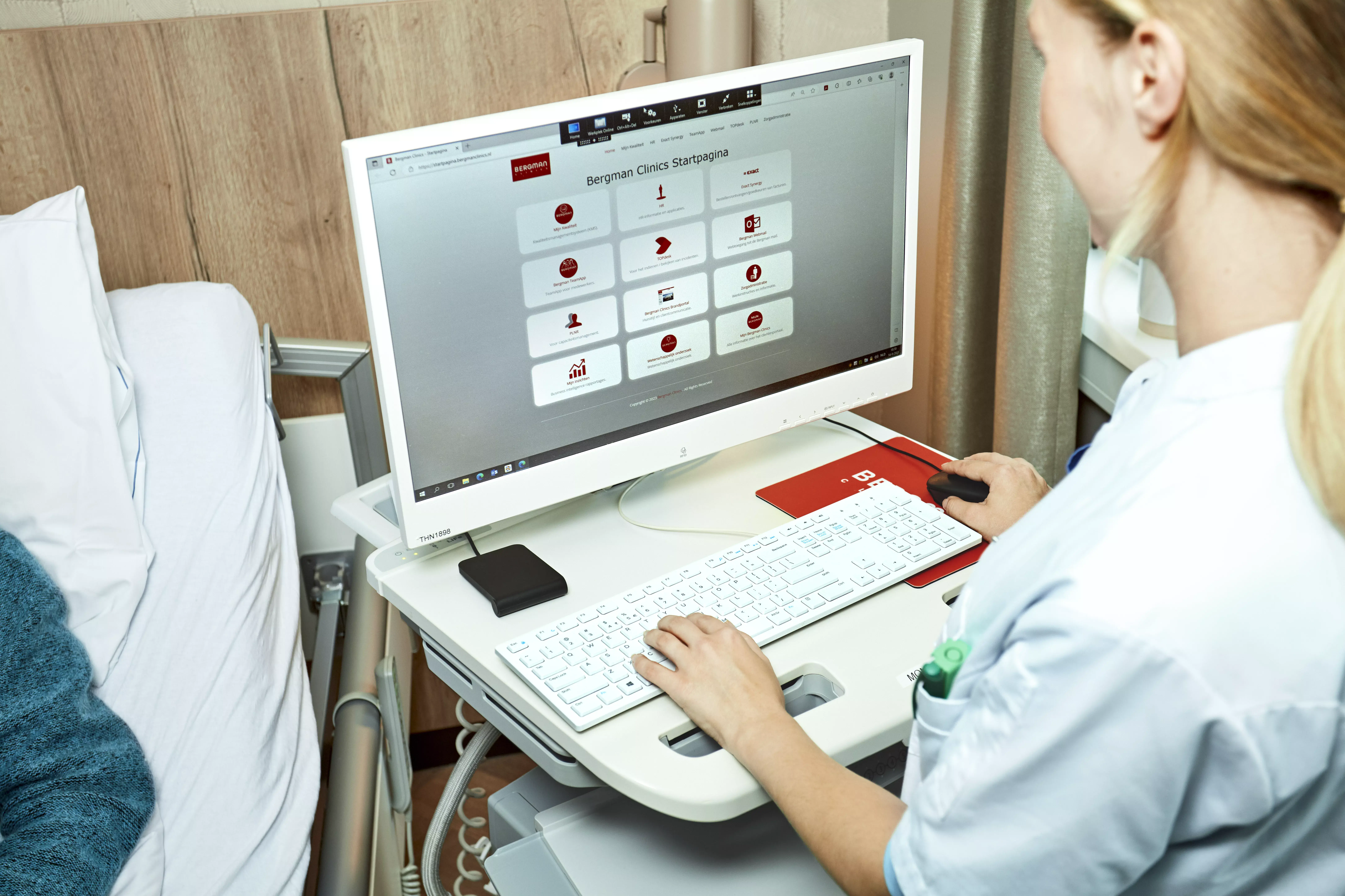 Een vrouwelijke arts zit naast een ziekenhuisbed achter een computer