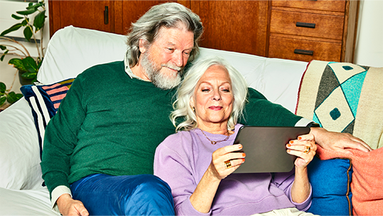 Stel met grijs haar ligt samen op de bank naar de tablet te kijken