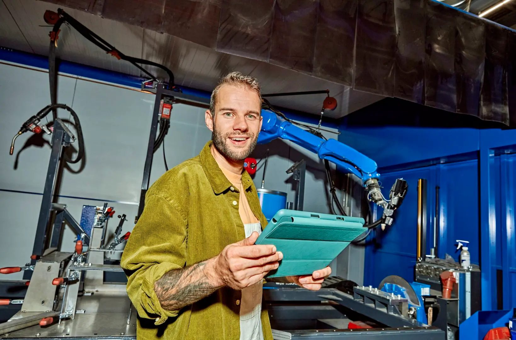 Man staat voor technische machines. Hij lacht naar de camera en houdt een tablet vast