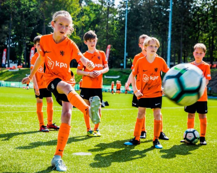 Een jeugdvoetbalteam op het veld in oranje shirts. Het meisje op de voorgrond trapt een bal weg.