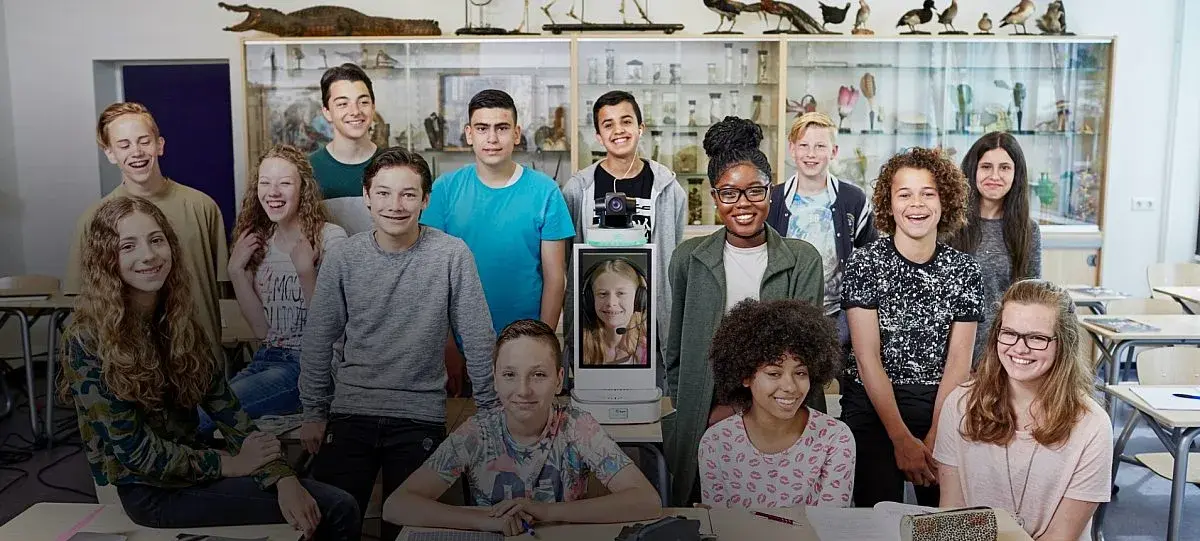 Leerling verschijnt via de laptop thuis op De Klasgenoot ICT-apparatuur in het klaslokaal en neemt zichtbaar deel tussen klasgenoten en leerkracht.