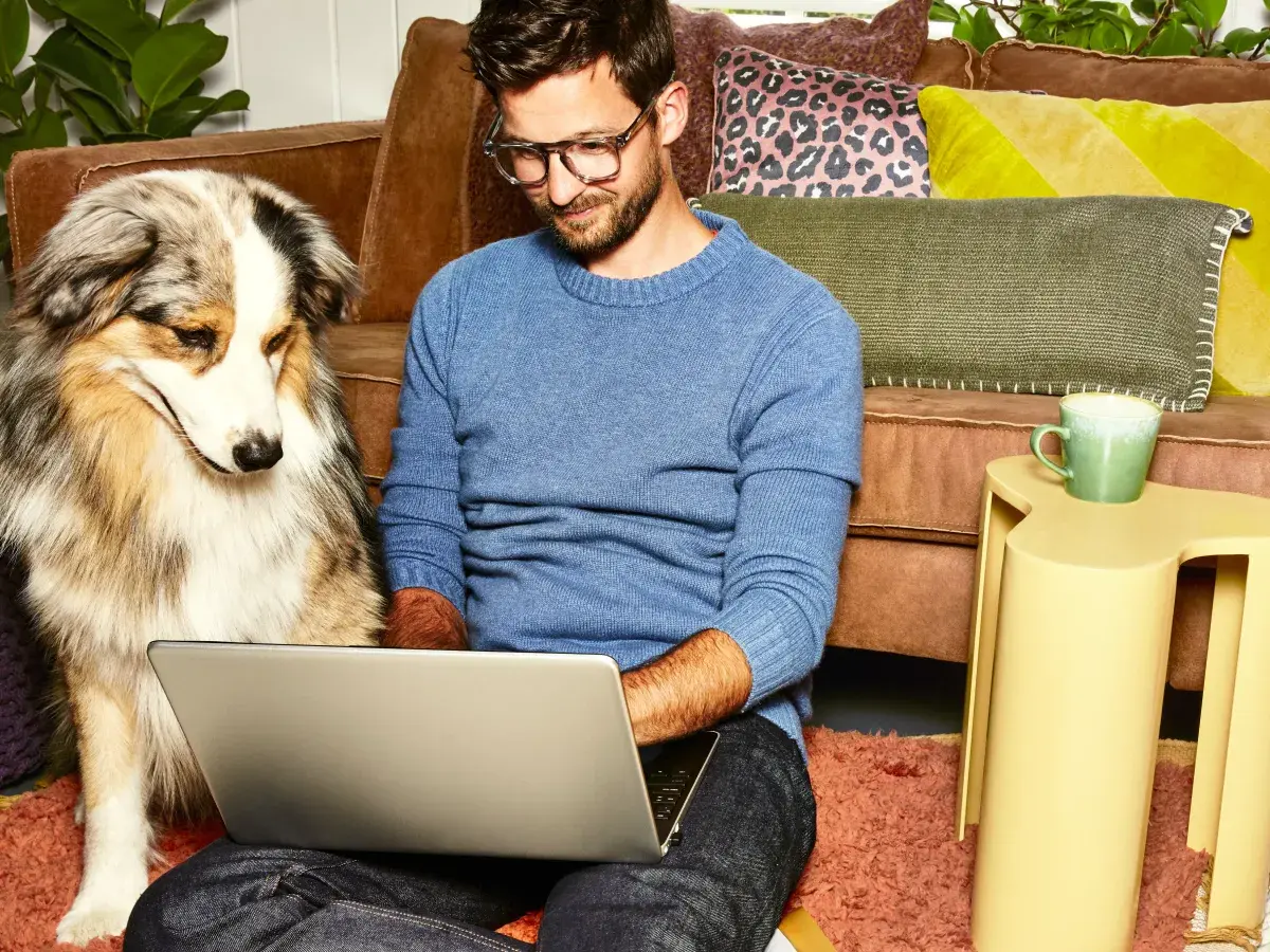 Een man zit op de grond voor een bank. Hij heeft zijn laptop op schoot. Naast hem zit een hond.