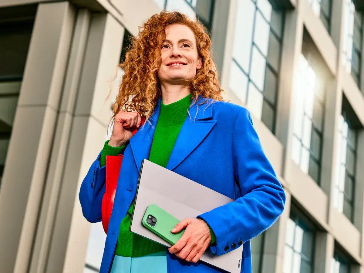 Een lachende vrouw loopt buiten. In haar hand heeft ze een telefoon en laptop vast.