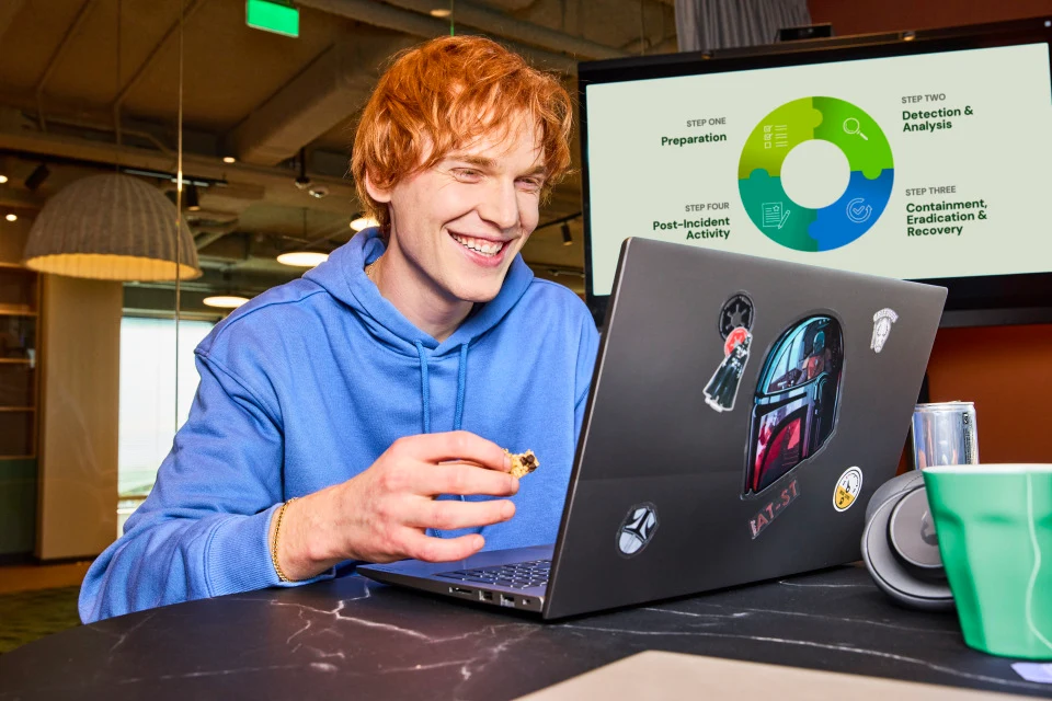 Jongen in blauwe trui achter laptop met stickers en een koekje in zijn hand